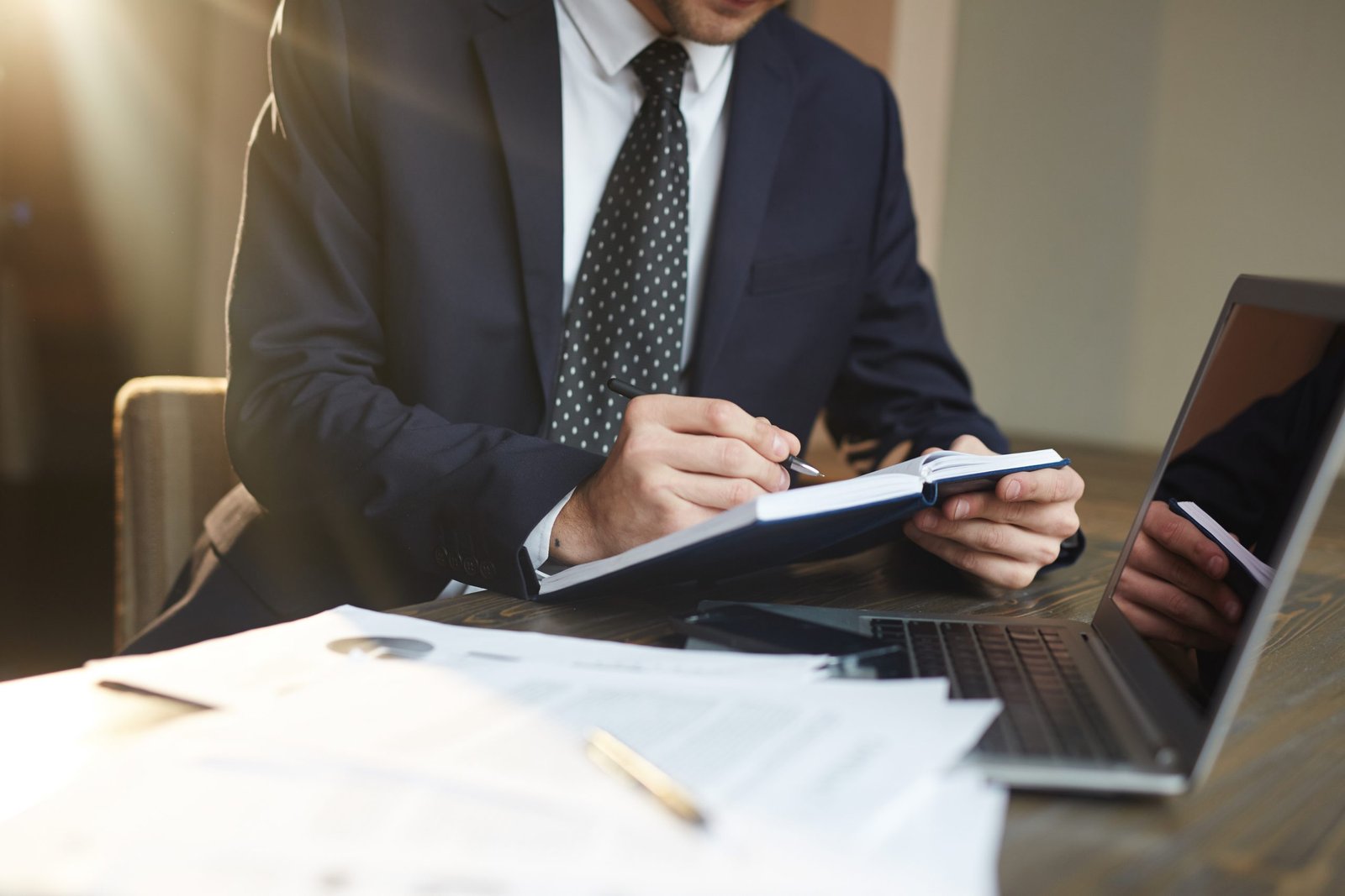 Closeup portrait of unrecognizable successful businessman wearing black formal suit  writing in planner while working with documentation and laptop